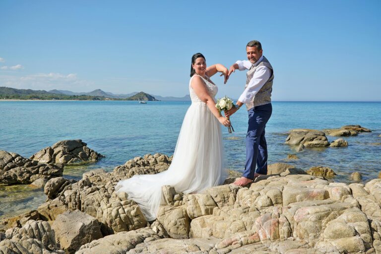 Brautpaar formt mit den Armen ein Herz vor dem atemberaubenden Meer, Strandhochzeit - meer sardinien urlaub