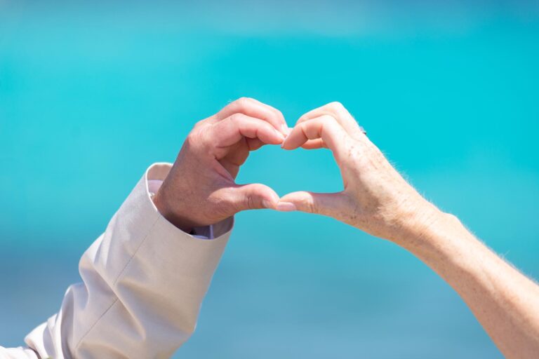 dei Hände des Brautpaares formen ein Herz vor einem azurblauen Meer, Strandhochzeit - meer sardinien urlaub
