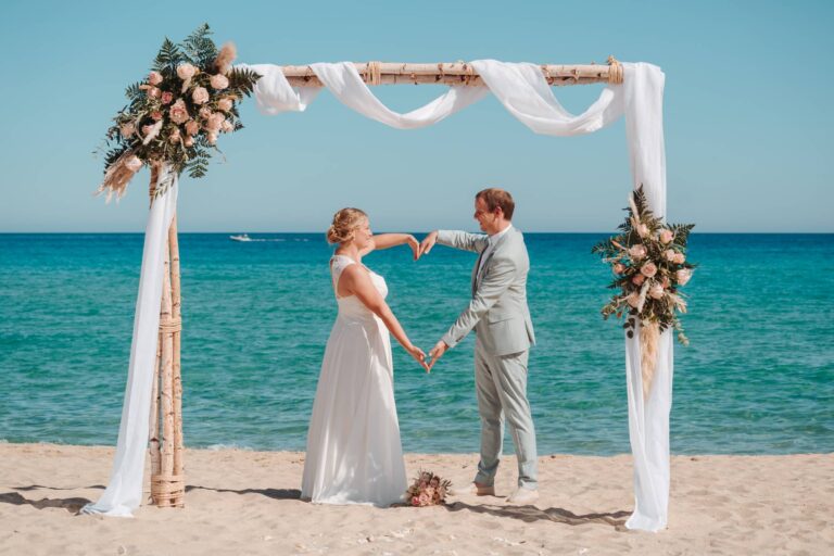 das Brautpaar formt ein großes Herz am Traubogen, Strandhochzeit - meer sardinien urlaub