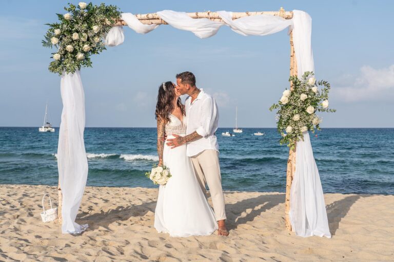 das glückliche Brautpaar unter dem weiß geschmückten Trauungsbogen, Strandhochzeit - meer sardinien urlaub