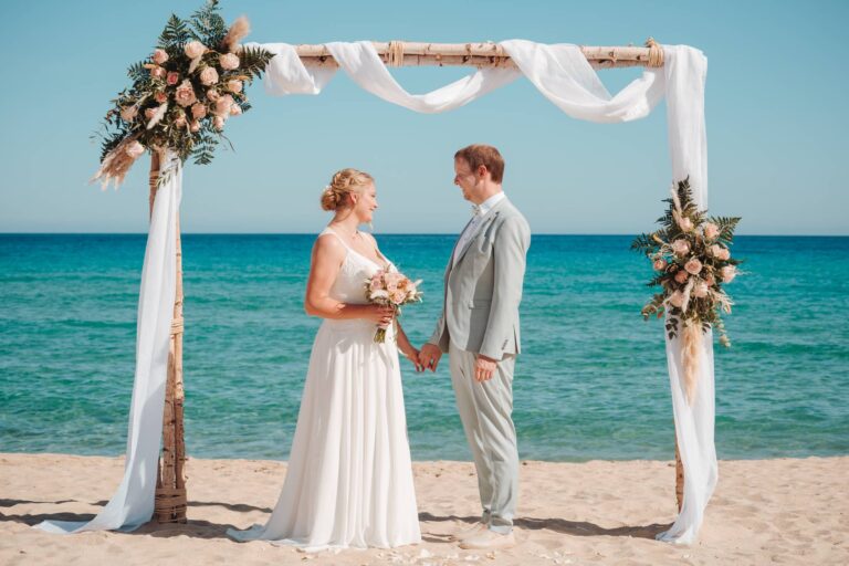 wahrlich glücklich lächelt sich das Hochzeitspaar unter dem Traubogen an, Strandhochzeit - meer sardinien urlaub