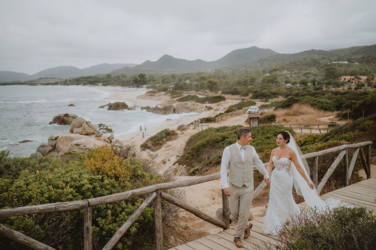 Brautpaar-auf-einem-Holzsteg-mit-der-Küste-im-Hintergrund,-Strandhochzeit---meer-sardinien-urlaub