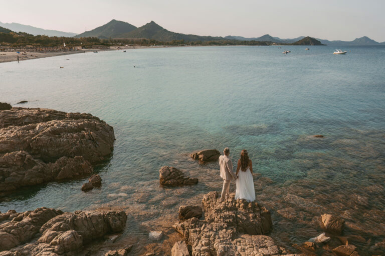 Brautpaar-blickt-andächtig-auf-den-Felsen-stehend-zum-Meer,-Strandhochzeit---meer-sardinien-urlaub
