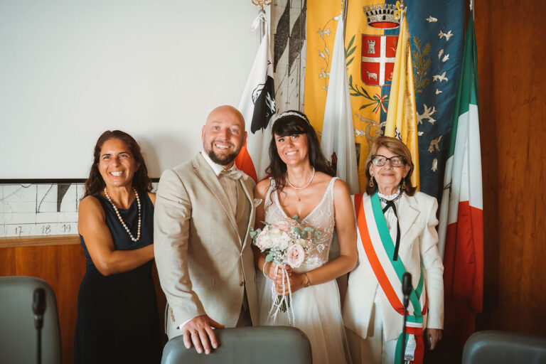 Erinnerungsfoto-des-Brautpaares-mit-der-Übersetzerin-und-der-Standesbeamtin-vor-den-Flaggen-im-Gemeindesaal,-Trauung-im-Standesamt---meer-sardinien-urlaub