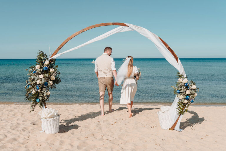Hand-in-Hand-geht-das-Brautpaar-hinter-dem-Traubogen-zum-Meer,-Strandhochzeit---meer-sardinien-urlaub
