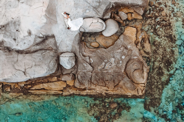 atemberaubende-Drohnen-Aufnahme-des-auf-einem-Felsen-liegenden-Brautpaares,-Strandhochzeit---meer-sardinien-urlaub