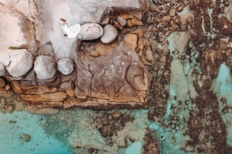 atemberaubende-Drohnen-Aufnahmen-des-auf-einem-Felsen-liegenden-Brautpaares,-Strandhochzeit---meer-sardinien-urlaub