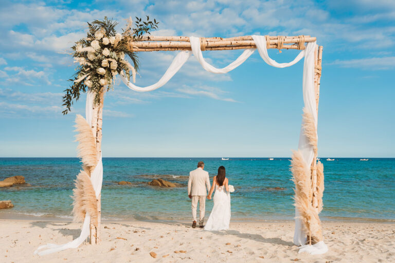 das-Brautpaar-spatziert-Richtung-Meer,-im-Vordergrund-der-harmonische-Traubogen,-Strandhochzeit---meer-sardinien-urlaub