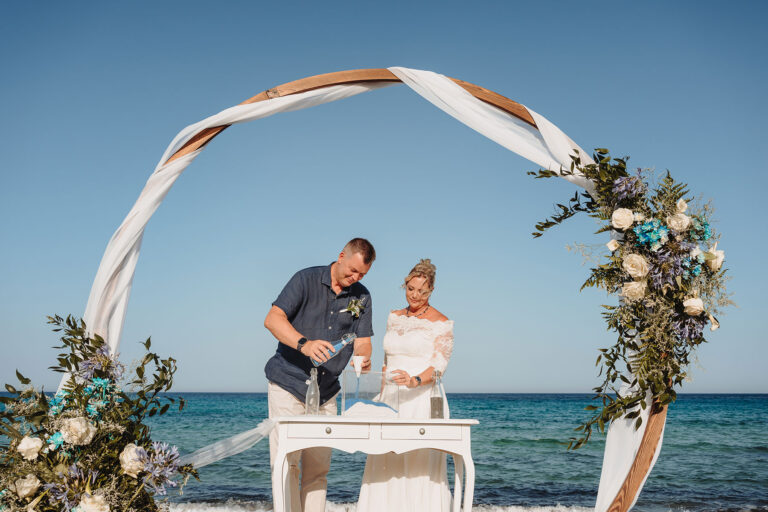 das-Brautpaar-vollführt-das-Sandritual-unter-dem-runden-Traubogen,-Strandhochzeit---meer-sardinien-urlaub