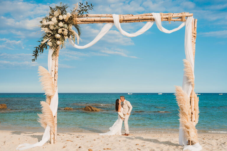 küssendes-Brautpaar-hinter-dem-Traubogen,-Strandhochzeit---meer-sardinien-urlaub