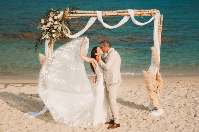 verspielt-hält-die-Braut-die-Schleppe-ihres-Spitzen-Kleides-hoch,-im-Hintergrund-der-Traubogen,-Strandhochzeit---meer-sardinien-urlaub