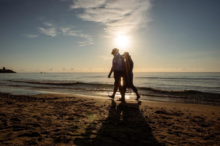 Braut-und-Bräutigam-spazieren-im-Gegenlicht-am-Meeresufer-nach-ihrer-Strandhochzeit---meer-sardinien-urlaub