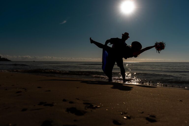 ausgelassen-posiert-das-Brautpaar-im-Gegenlicht-vor-der-aufgehenden-Sonne,-Strandhochzeit---meer-sardinien-urlaub