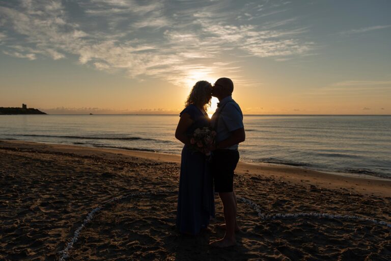 gefühlvoll-küsst-der-Bräutigam-die-Braut-auf-die-Stirn-vor-einer-aufgehenden-Sonne-am-Strand,-Strandhochzeit---meer-sardinien-urlaub