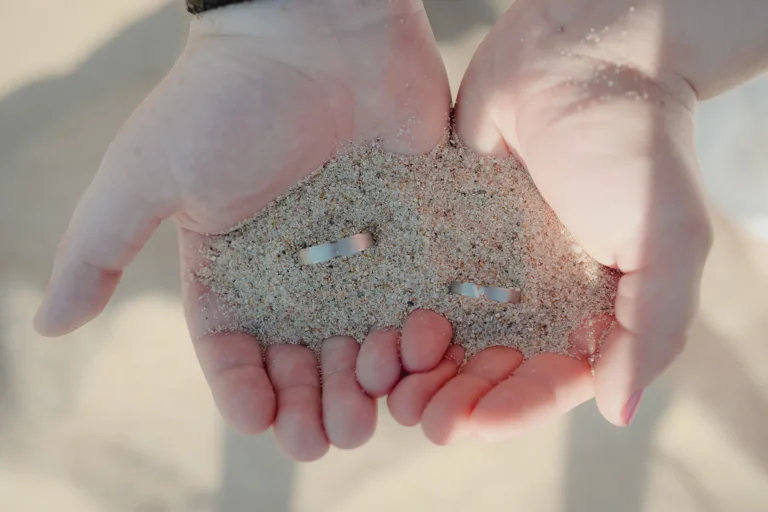 die Hände des Brautpaares halten die Eheringe mit Sand, Strandhochzeit - meer sardinien urlaub